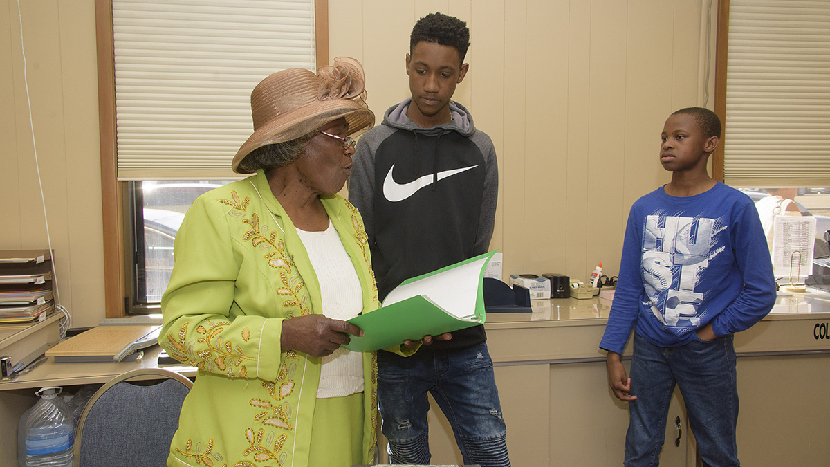 A young boy stands next to and looks down at a woman wearing a large brown hat and a bright green jacket.