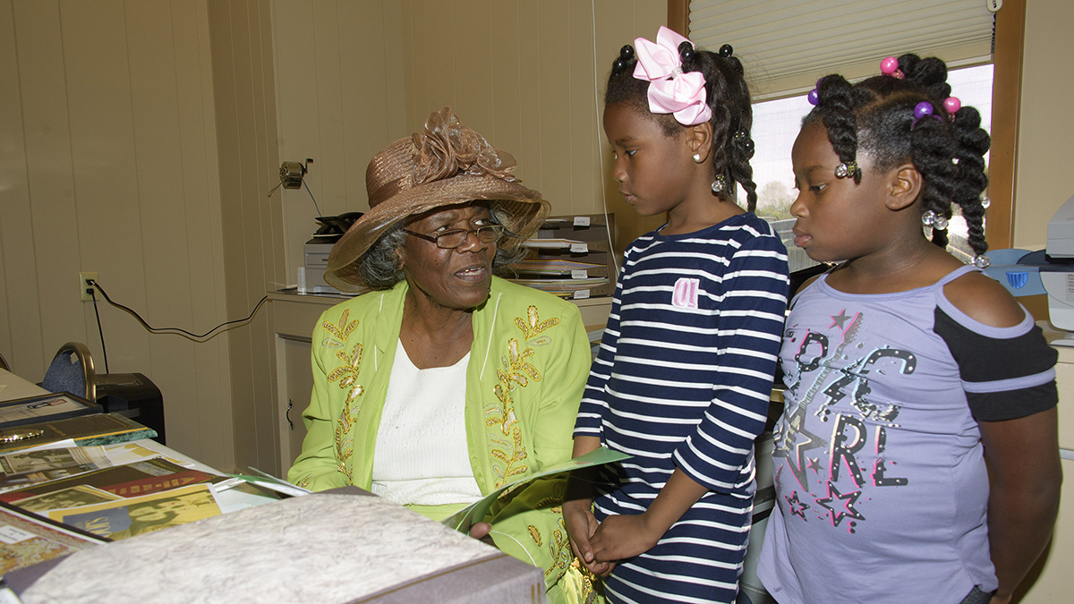 Two young girls and an older woman with a large brown hat and a bright green jacket talk to each other.