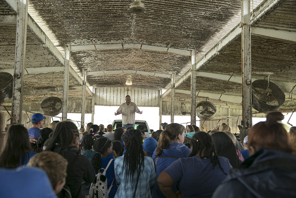 An adult male stands in the bed of a white pickup to address a crowd gathered in an open-sided dairy barn. 