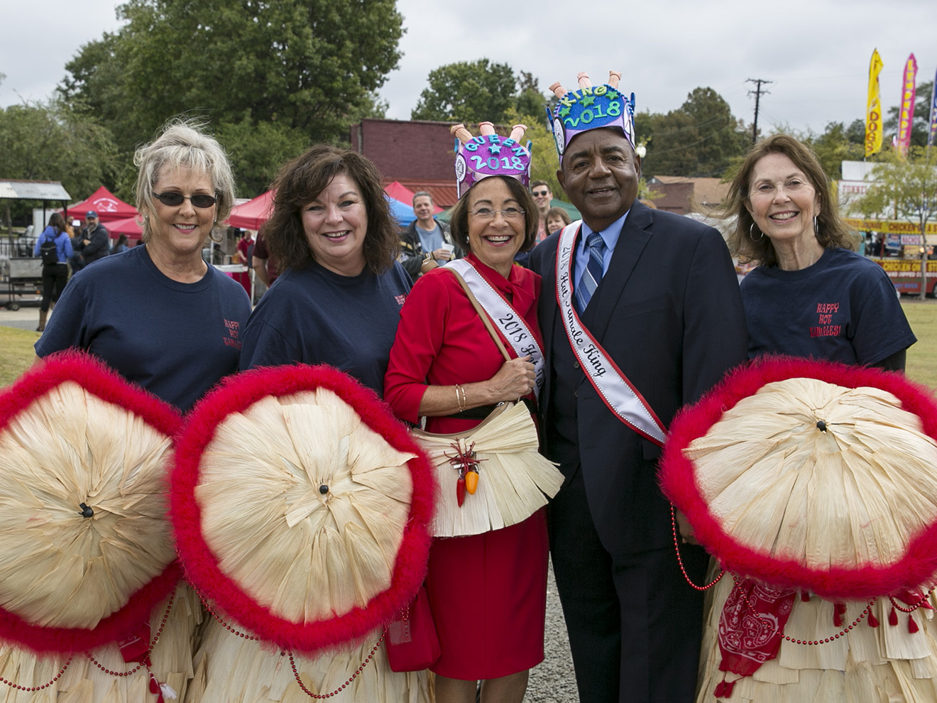 Three women holding red-rimmed straw stand on either side of a man and woman wearing crowns and white sashes. 