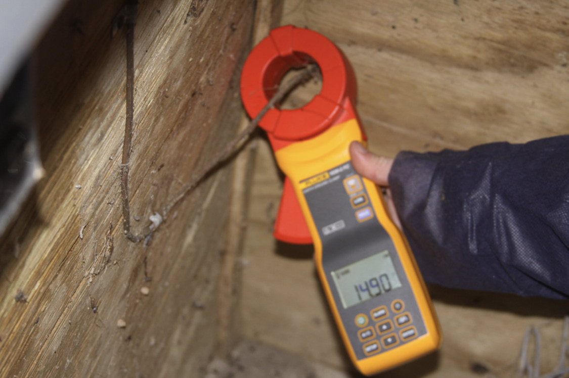 A person holds a ground resistance meter that shows a reading of 14.90 ohms.