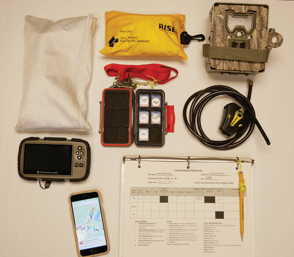 A layout of items for camera kits: a zipped bag of miscellaneous tools, SD card reader, smartphone displaying a Google map of camera sites, first aid kit in a yellow bag, SD card case with lanyard, open binder showing station field forms with an attached mechanical pencil, camouflage assembled camera trap, and cable lock with cable.