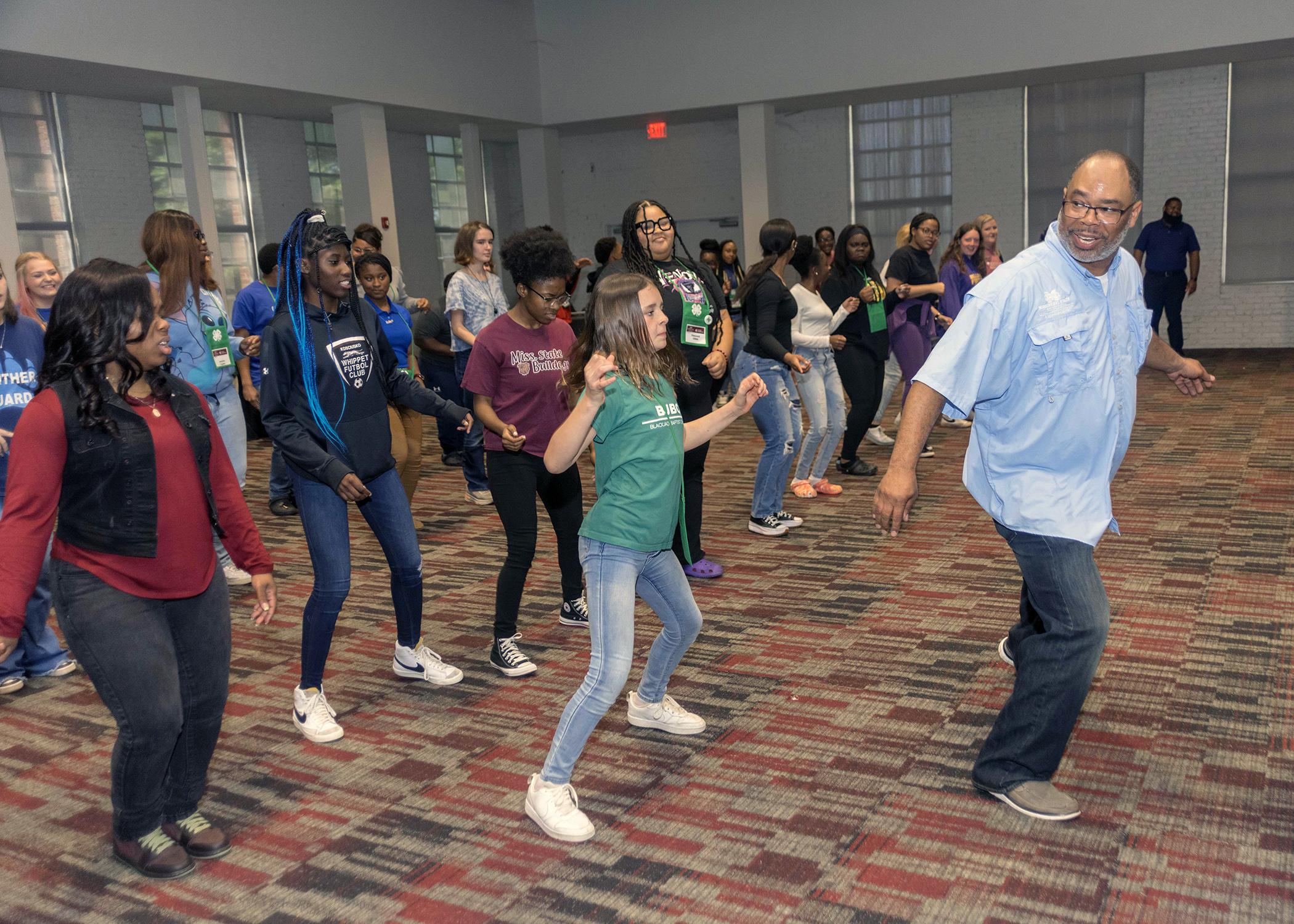 Man leads room full of teenagers in dance exercise.