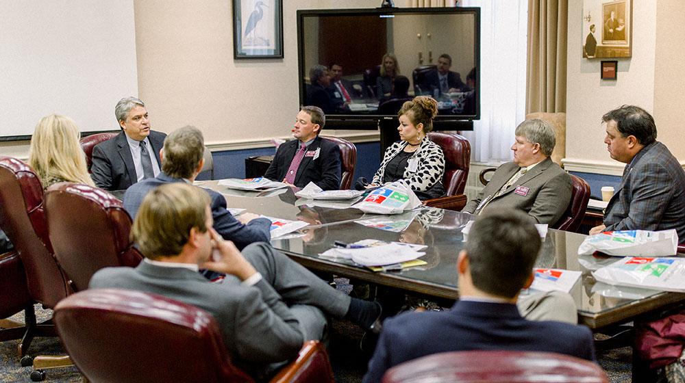 Group of people seated at boardroom table, listening to speaker.