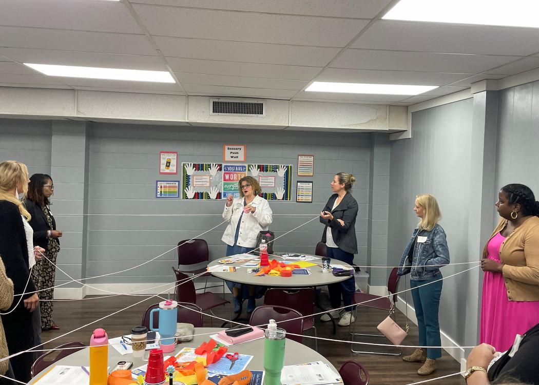 A group in a circle holds a white string that crisscrosses a classroom.