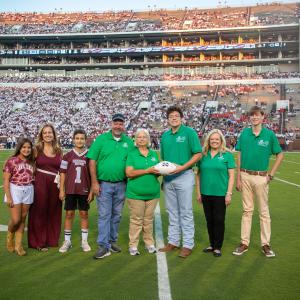 A group of people standing on a football field; the woman and man in the center holding a white football.