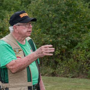 A man wearing a vest and green shirt, talking and gesturing.