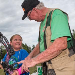 A man wearing a vest and green shirt, talking to a little girl smiling and holding an archery bow.