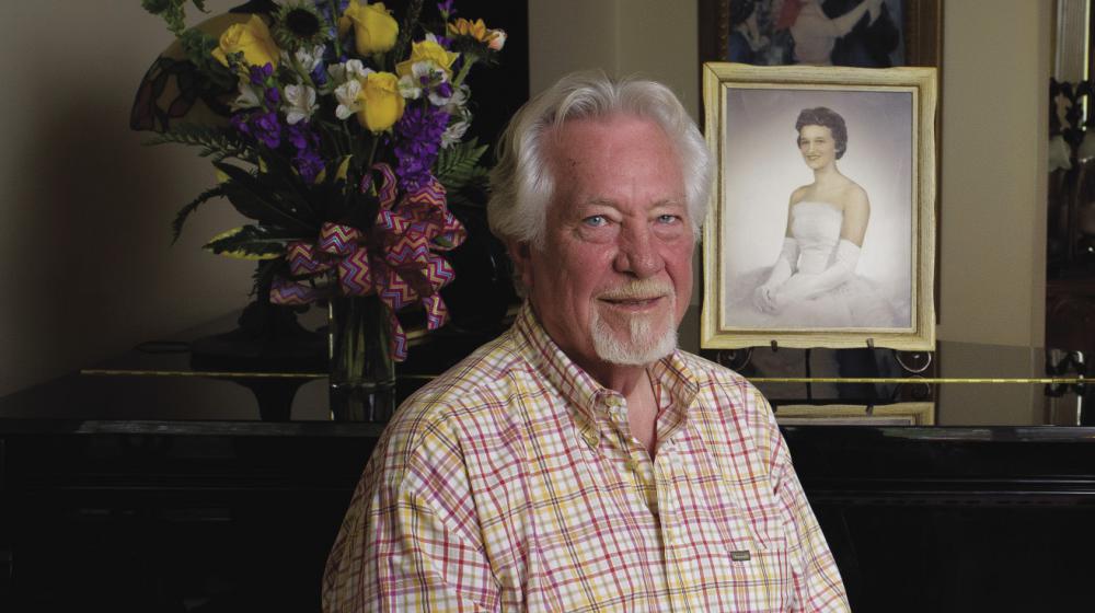 A male retiree in a plaid shirt and khaki slacks sits in front of a piano. On its soundboard is a picture of his late wife in her wedding gown and gloves.