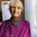 An smiling elderly woman stands next to a window. 