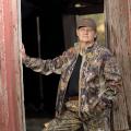 A woman wearing a camouflage jacket and pants rests her hand on the entrance to an old wooden building. 