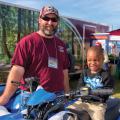 A man stands next to an ATV with a young boy sitting on the seat.