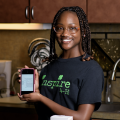 A smiling teenage girl stands behind a table covered in ingredients for a recipe displayed on a phone screen she holds in front of her. 