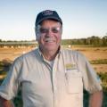 A smiling man with a "dedicated to dairy" logo on his shirt, stands in a pasture.