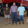 Three men and a dog stand in front of farm equipment.