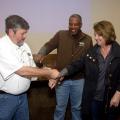 A man mimics removing a fishing hook from a woman’s arm during a safety demonstration as another man looks on.