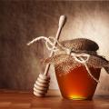 Wooden honey dipper lying against a small jar of honey that is covered with a decorative piece of fabric tied with a string.