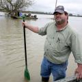 A man in work clothes, baseball cap and wading boots stands in water outside his boat holding a paddle.