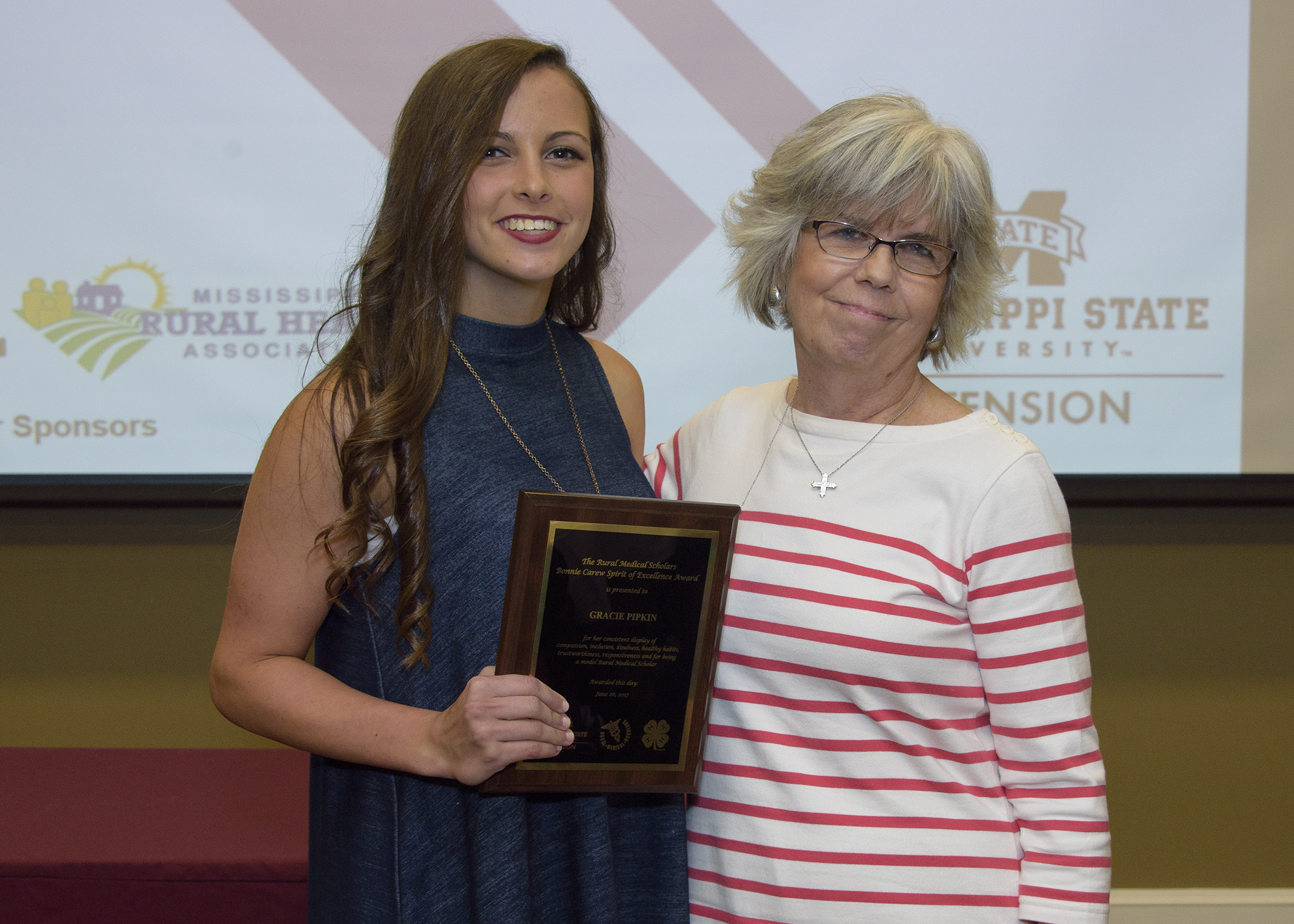 brunette female holds award plaque standing next to another female.